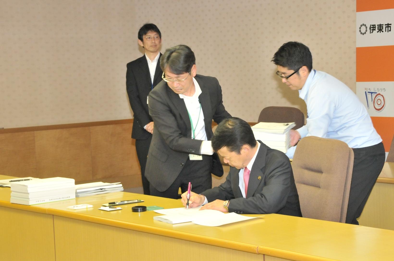 2017年5月29日書類の束を運ぶ市職員の側で書類へサインをする市長の写真