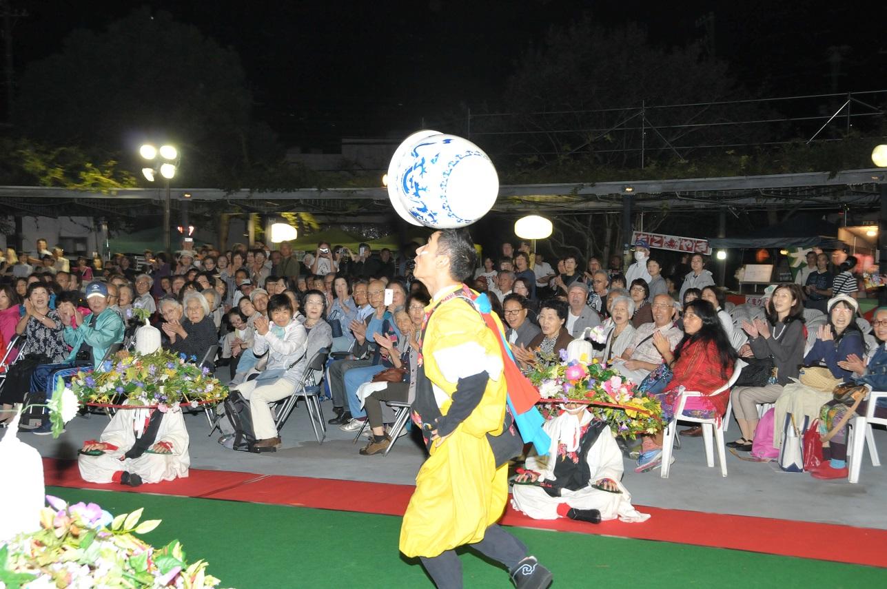壺を頭に乗せて歩くパフォーマンスをする男性と席に座り拍手をする観客の写真