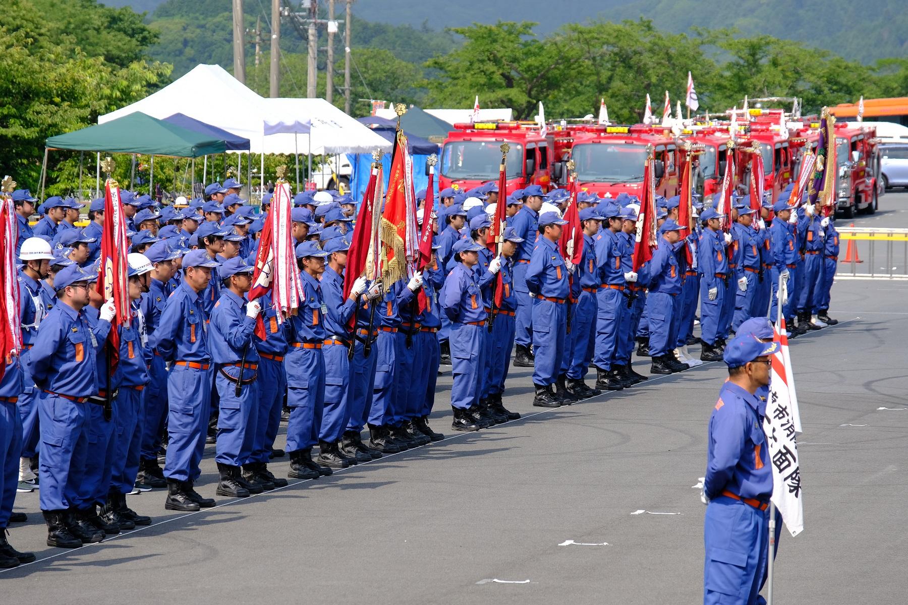 並ぶ消防車と消防団旗を持ち整列する消防団員の写真